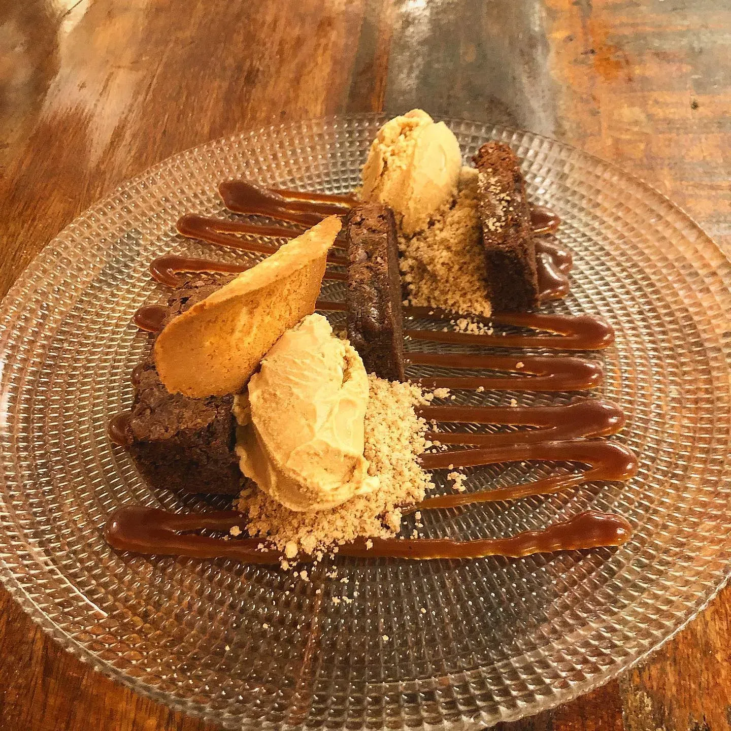 Postre de brownie con helado, llovizna de caramelo y una oblea sobre un plato de vidrio.