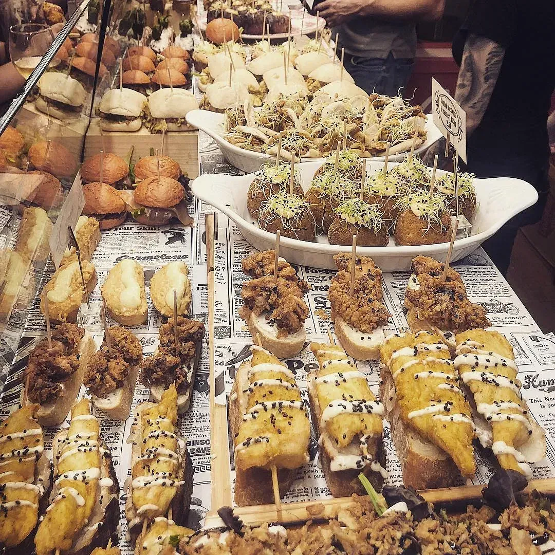 Surtido de tapas españolas expuestas en un mostrador, incluidos sándwiches, brochetas y croquetas.