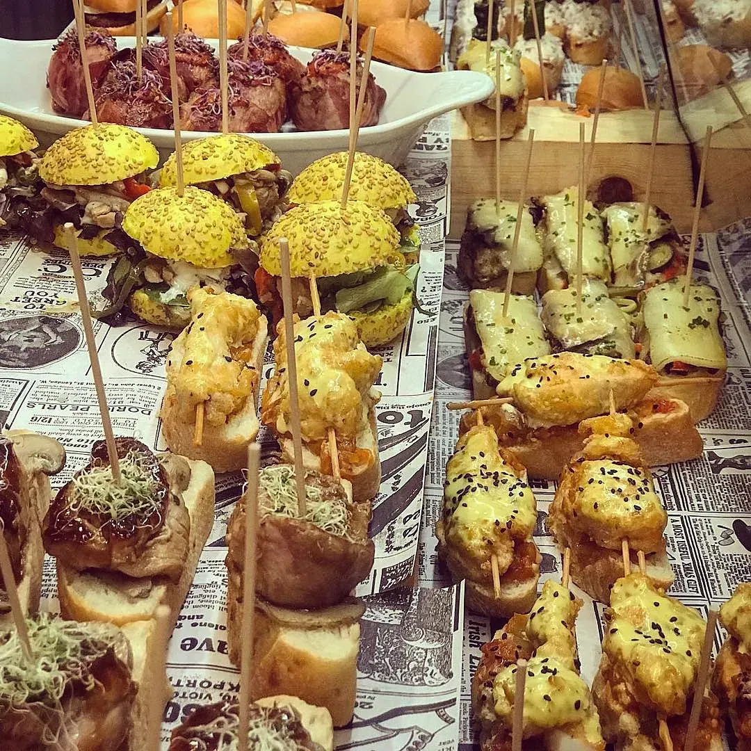 Tapas variadas sobre un mostrador, incluyendo minihamburguesas, brochetas y sándwiches abiertos, en papel de periódico.