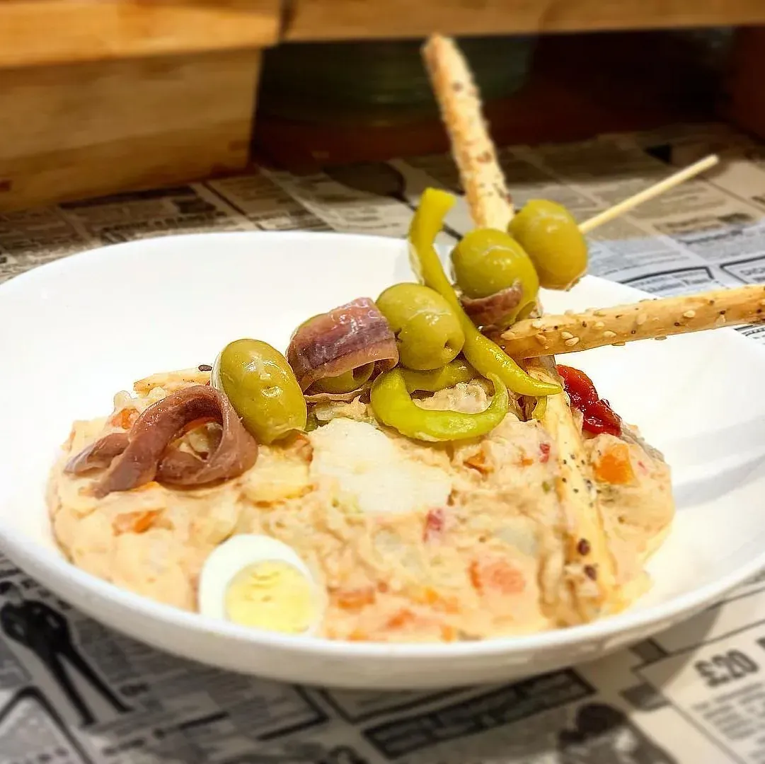 Ensalada de patatas con aceitunas, anchoas, pimientos y palitos de pan en un bol blanco.