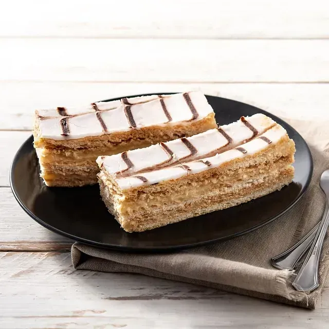 Dos pasteles de capas con glaseado blanco y rayas de chocolate negro sobre un plato negro, con una servilleta y cubiertos sobre una superficie de madera blanca.