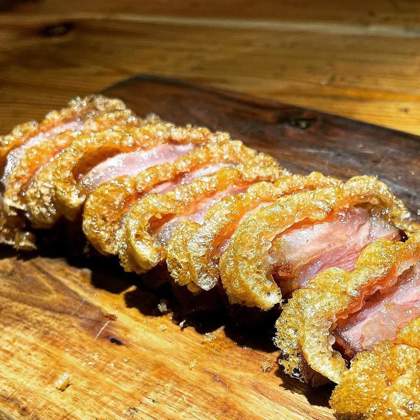 Panceta de cerdo crujiente cortada en rodajas sobre una tabla de madera. Piel dorada, carne rosada visible.