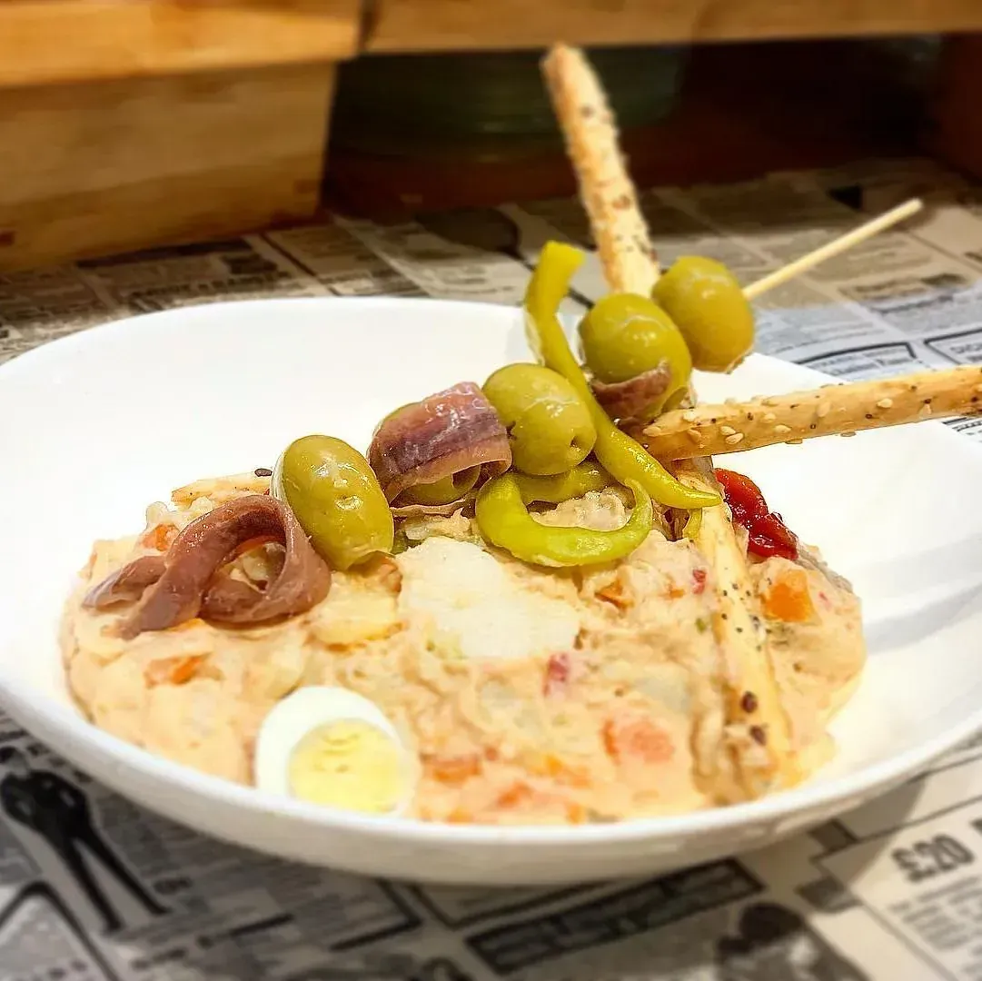 Ensalada de patatas, aceitunas, anchoas, pimiento, huevo, palitos de pan, en un bol blanco sobre un periódico.