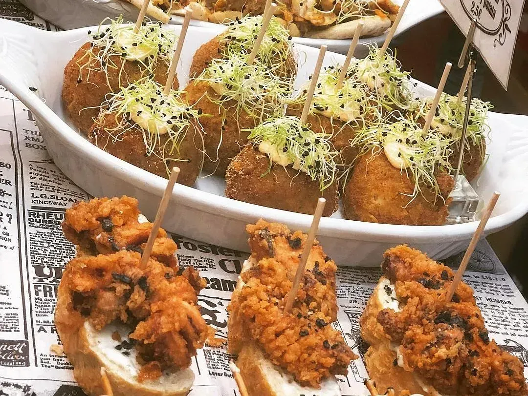 Tapas: pan cubierto con frituras y croquetas en un plato blanco, decorado con palillos y brotes.