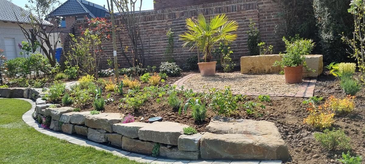 Ein Garten mit vielen Pflanzen und Steinen vor einer Backsteinmauer.