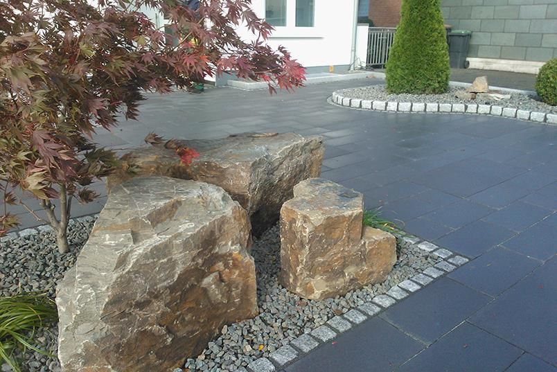Ein großer Stein liegt mitten auf einer Einfahrt neben einem Baum.