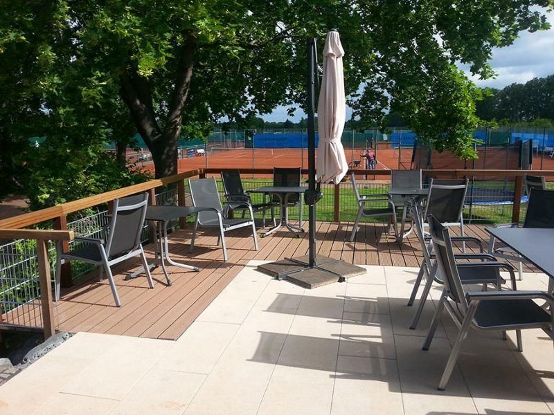 Eine Terrasse mit Tischen und Stühlen und einem Sonnenschirm mit einem Tennisplatz im Hintergrund