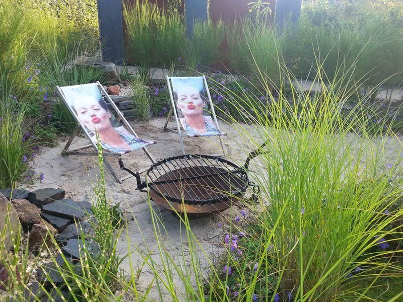 Zwei Gartenstühle mit einem Frauengesicht darauf stehen neben einer Feuerstelle.