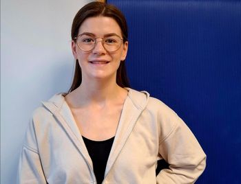 Eine Frau mit Brille und Kapuzenpullover steht vor einer blauen Wand.