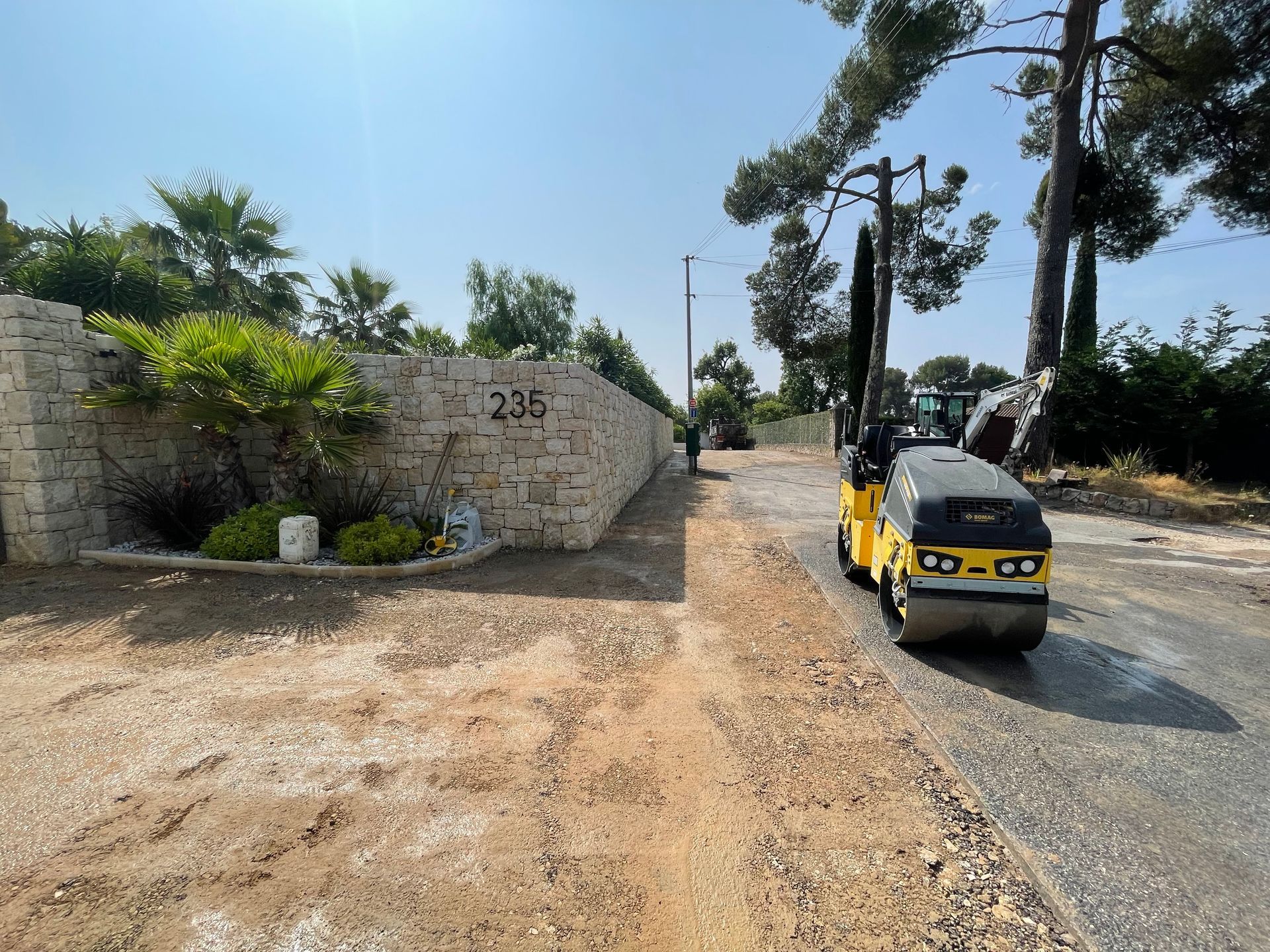 Rouleau compresseur compactant une allée en asphalte à côté d'un mur de pierre.