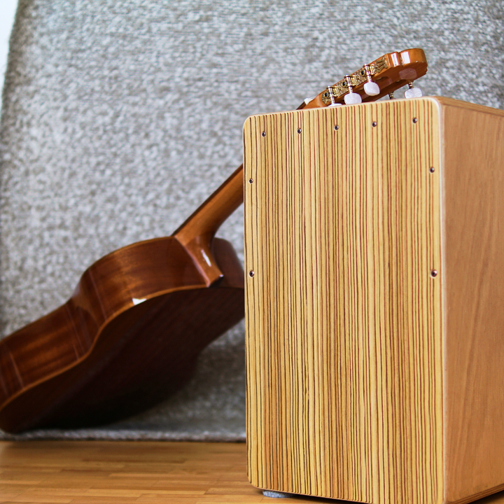 Cajón de madera junto a una guitarra clásica, apoyado sobre una alfombra gris sobre un piso de madera.
