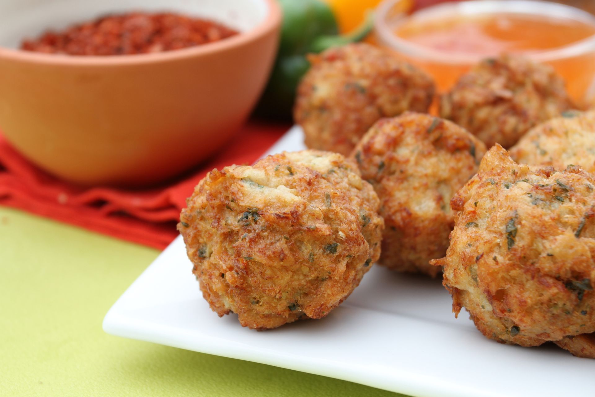 Des amuse-bouches frits, servis sur une assiette blanche avec deux petits bols de sauces à tremper sur une surface vert clair