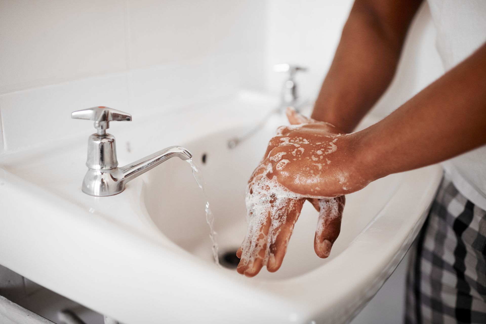 Une personne se lave les mains avec du savon et se les rince avec de l'eau courante sous un robinet argenté dans un lavabo blanc.