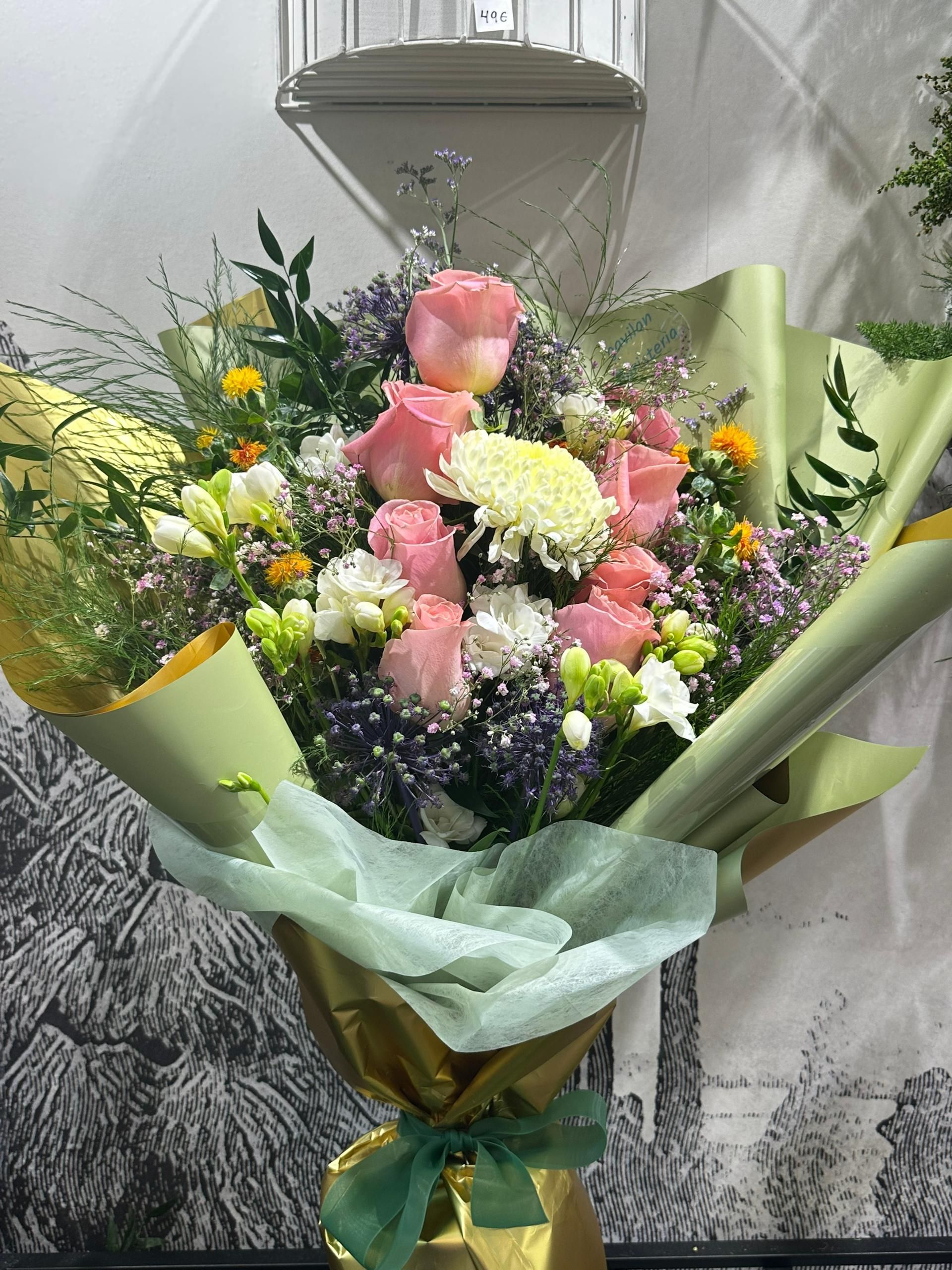 Ramo de rosas rosadas y flores variadas envuelto en papel verde y atado con una cinta dorada.