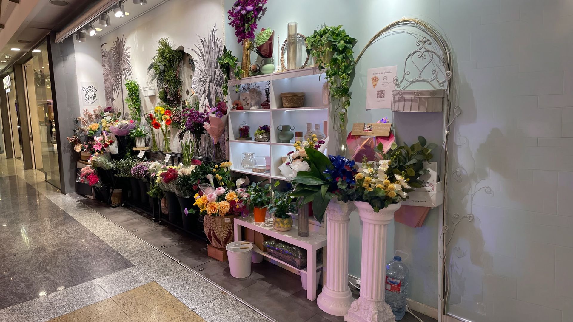 Exhibición de floristería decorada con coloridos ramos, estantes blancos y pedestales rosas a lo largo de un pasillo.