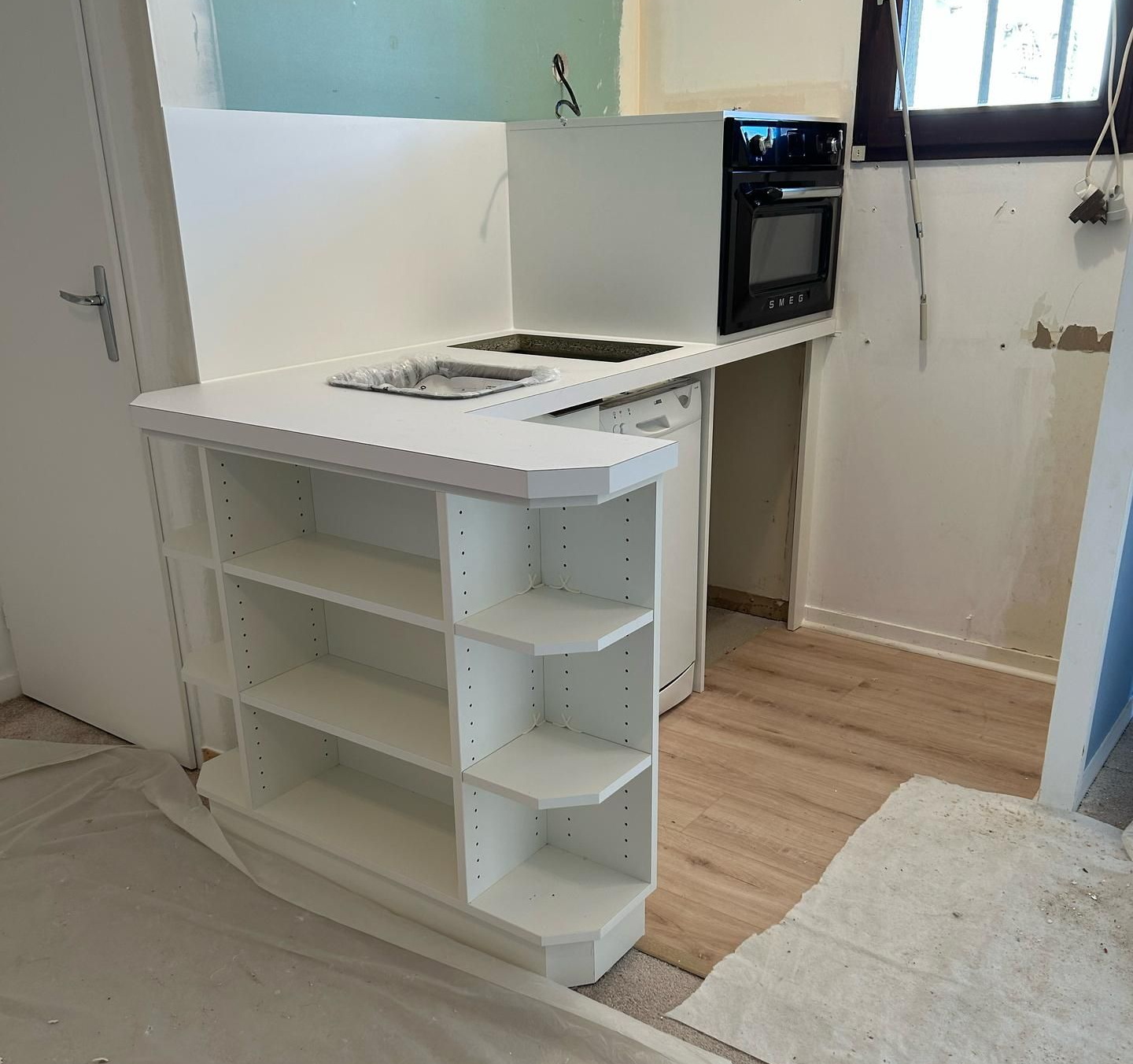 Un comptoir de cuisine blanc en forme de L avec des étagères ouvertes, un évier et un four encastré, lors de la rénovation d'une pièce.