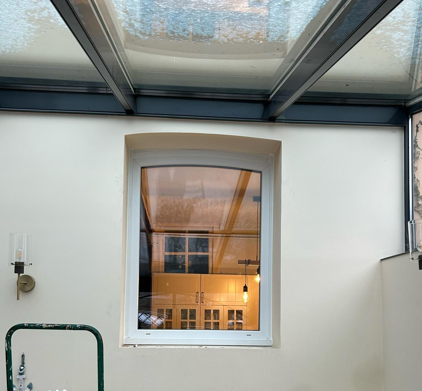 Une fenêtre à sommet incurvé, encastrée dans un mur blanc sous un panneau de plafond en verre, laisse apparaître le reflet d'une pièce intérieure.