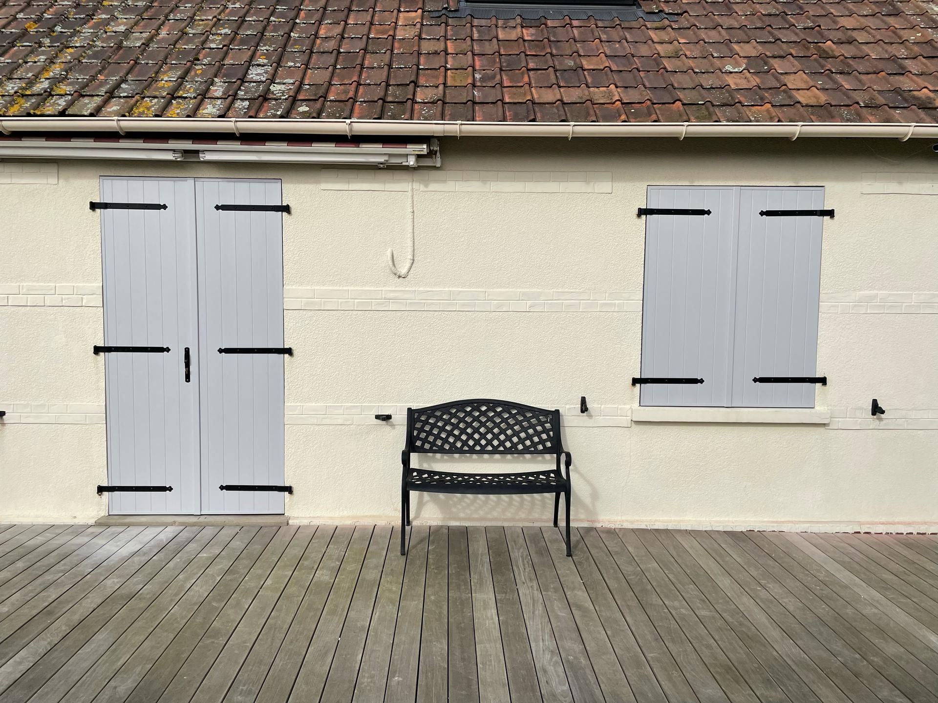 Un banc de jardin noir est installé sur une terrasse en bois devant un bâtiment aux portes et volets gris clair.