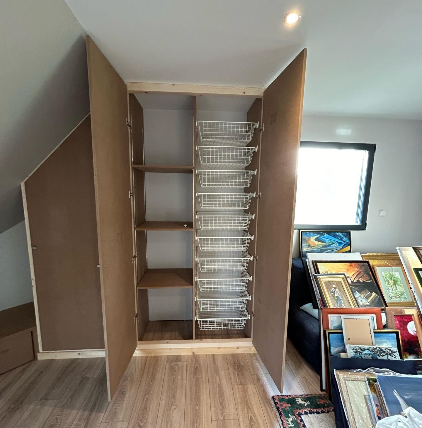 Un placard en bois sur mesure aménagé dans une pièce mansardée, comprenant des étagères et une colonne de paniers coulissants en fil de fer blanc.