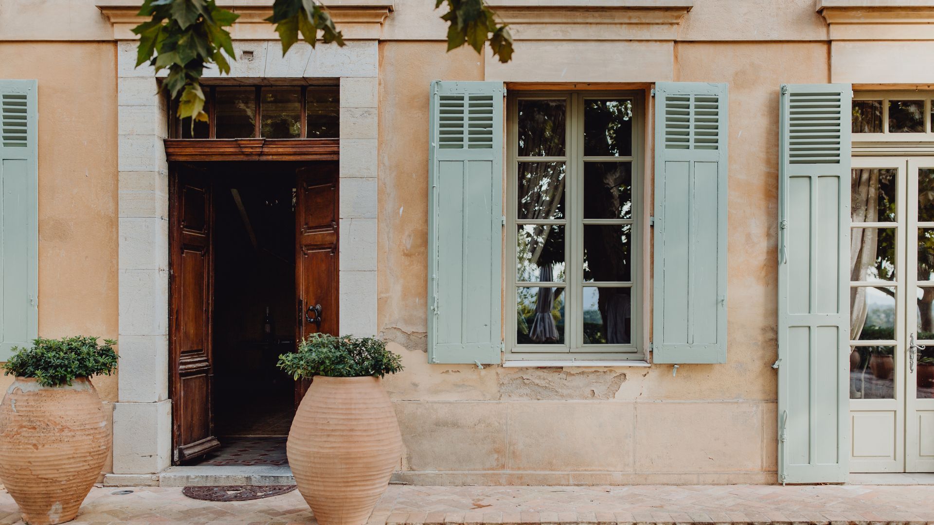 Une maison beige de style méditerranéen avec des volets vert clair, une porte en bois ouverte et deux grands pots en terre cuite.