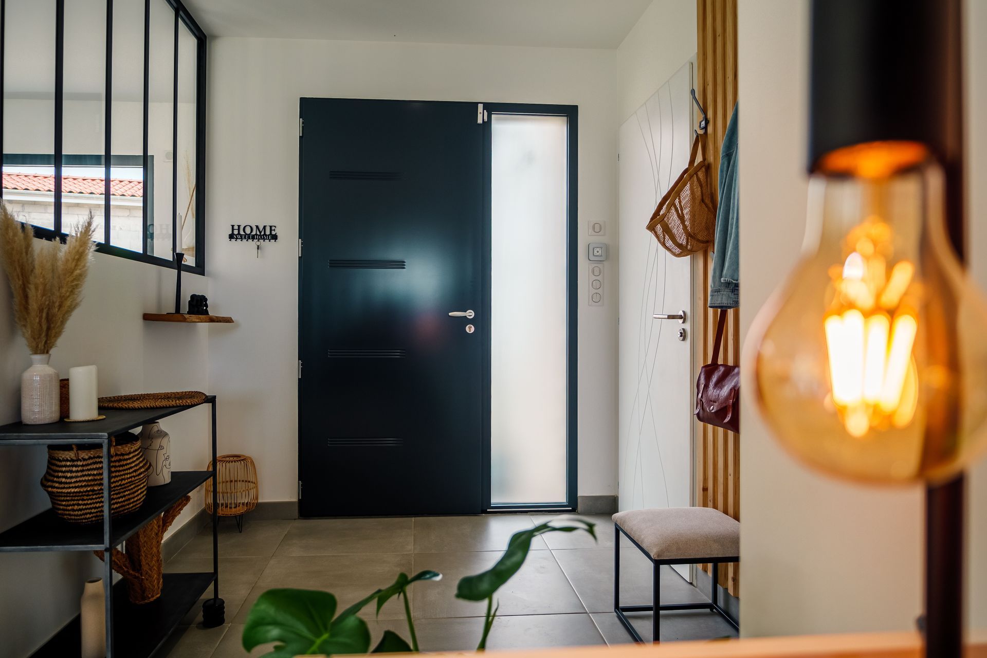 Une entrée moderne avec une porte noire mate, un panneau latéral en verre dépoli, une étagère et une ampoule au premier plan.