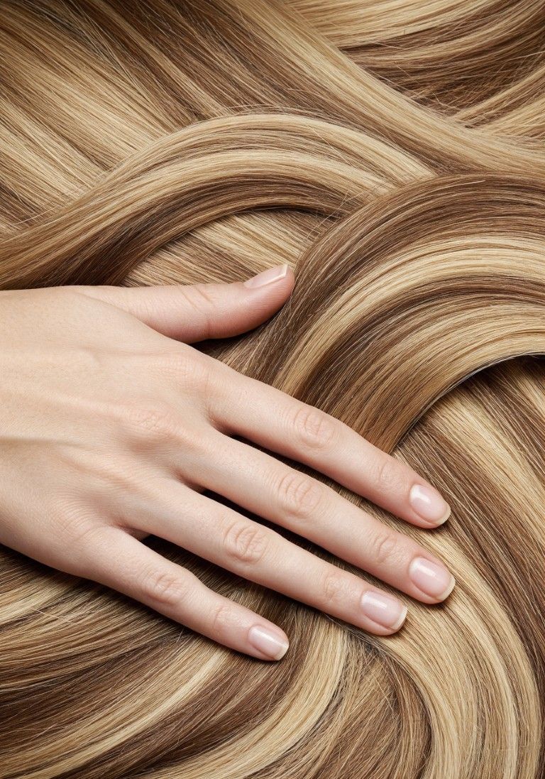Mano con uñas largas descansando sobre cabello rubio ondulado, mostrando mechones suaves con reflejos.