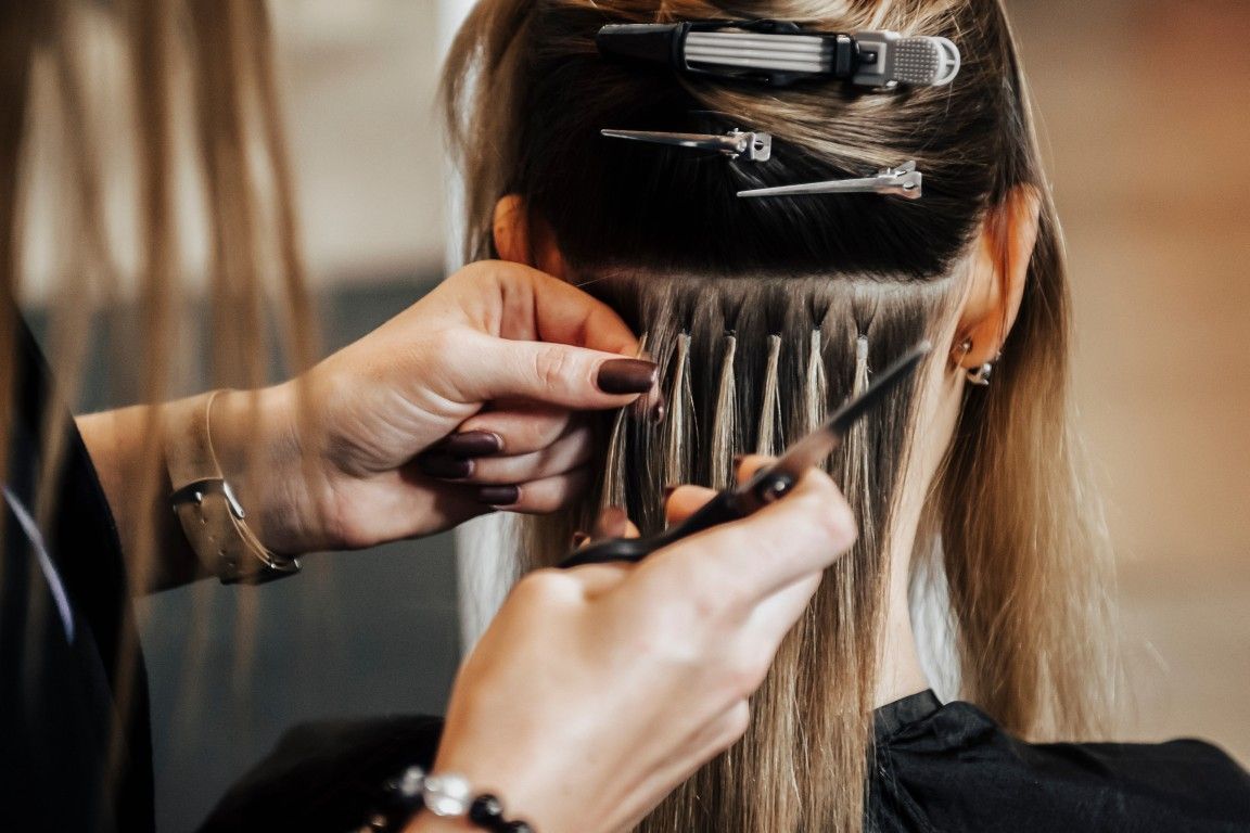 Una estilista aplica extensiones de cabello rubio con clips al cabello castaño de una clienta en una peluquería.