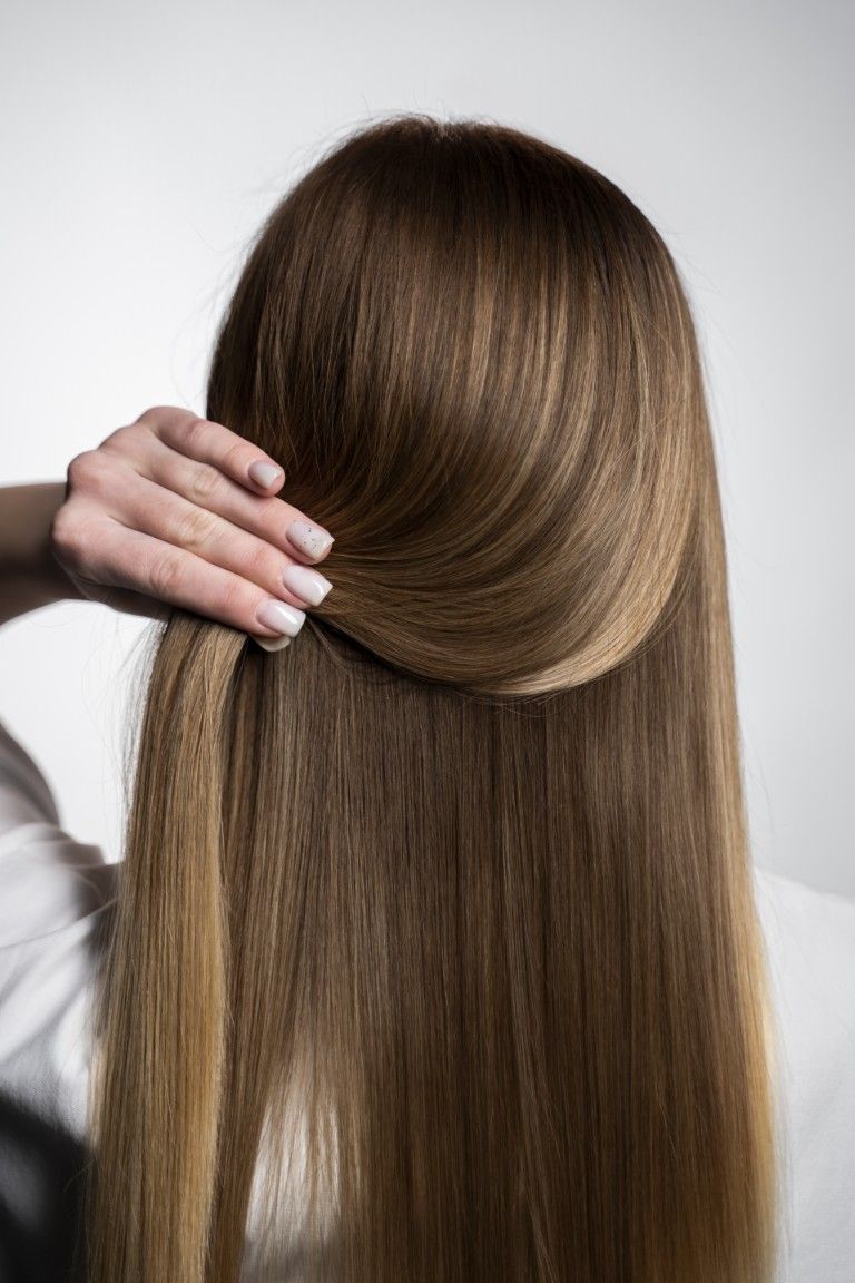 Levantando con la mano una sección de cabello largo, liso y castaño sobre un fondo claro.