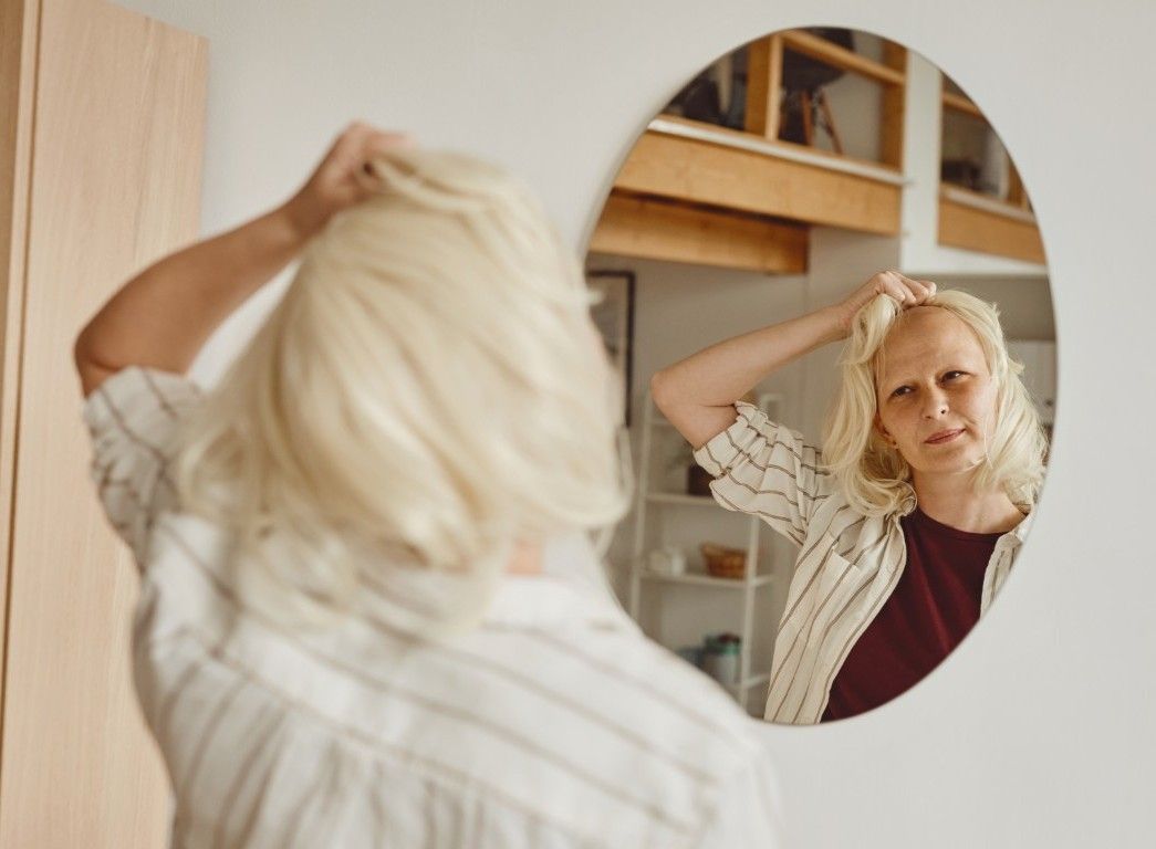 Una persona se arregla el cabello rubio mientras se mira en un espejo ovalado en una habitación de color claro.