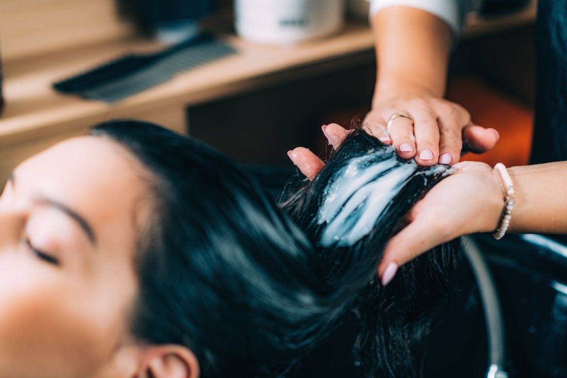 Una peluquera aplica champú mediante un masaje en el cabello oscuro de una clienta en el lavabo de la peluquería.