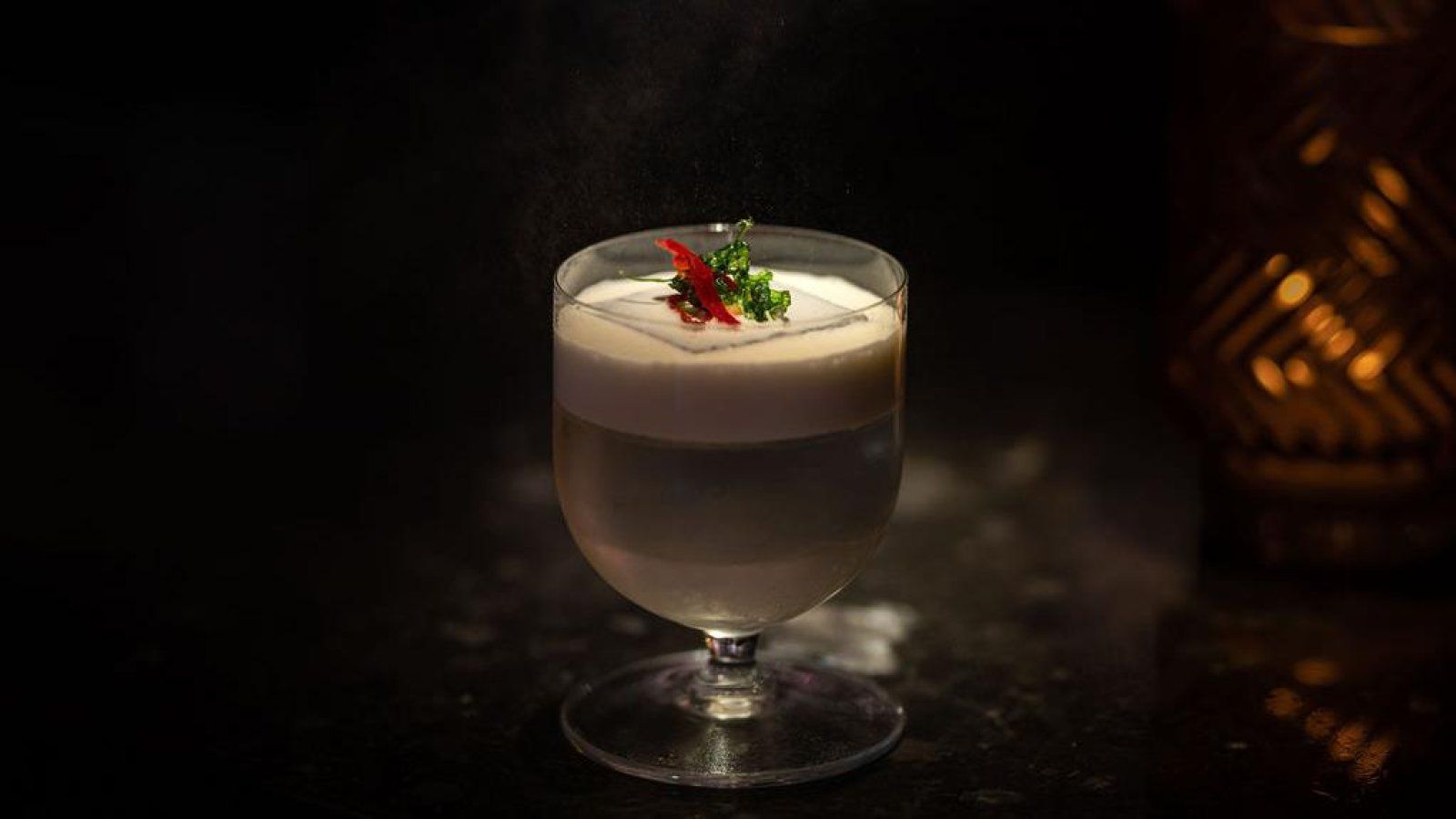 Cocktail with a frothy white top, garnished with red pepper and parsley. Set on a dark surface with a dimly lit background.