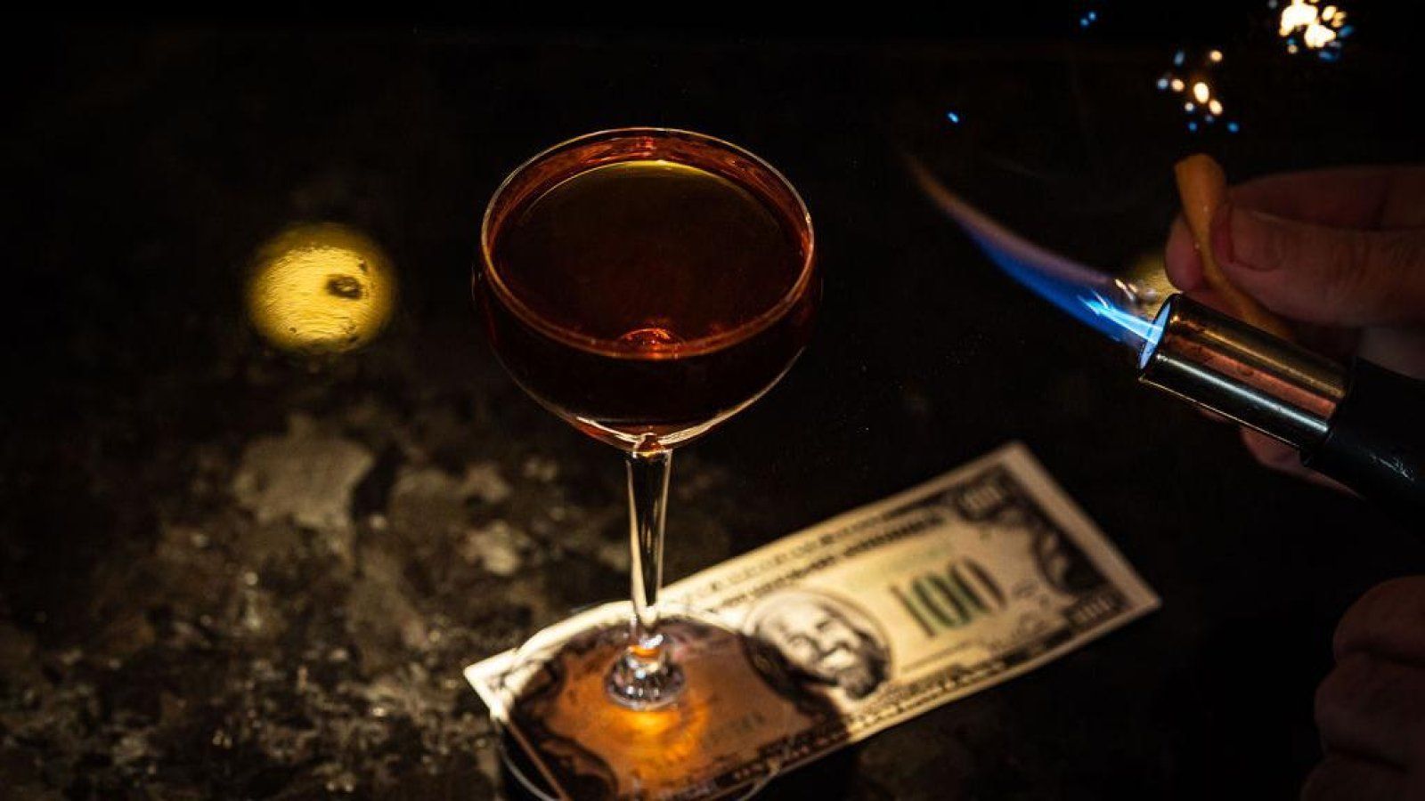 A person is lighting a lighter next to a glass of wine and a dollar bill.