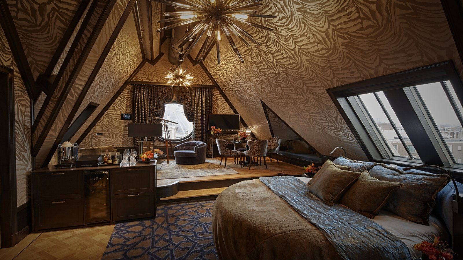 Hotel suite with slanted ceiling, circular bed, and seating area. Rich, dark tones and textured walls create a luxurious atmosphere.