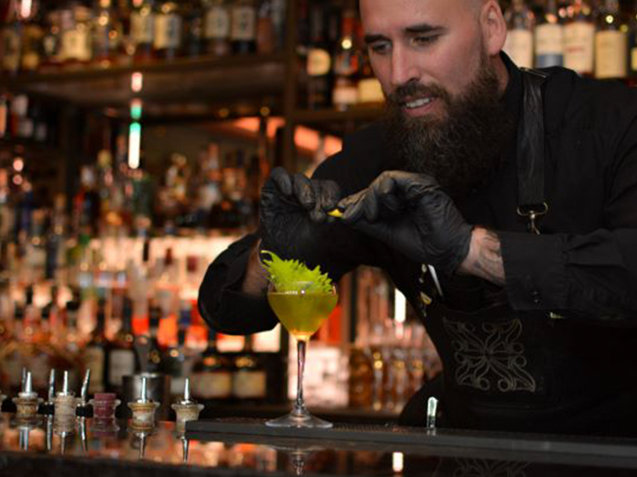 Een barman met een volle baard en zwarte handschoenen garneert een cocktail met een citrustwist. Hij staat achter een goed gevulde bar.