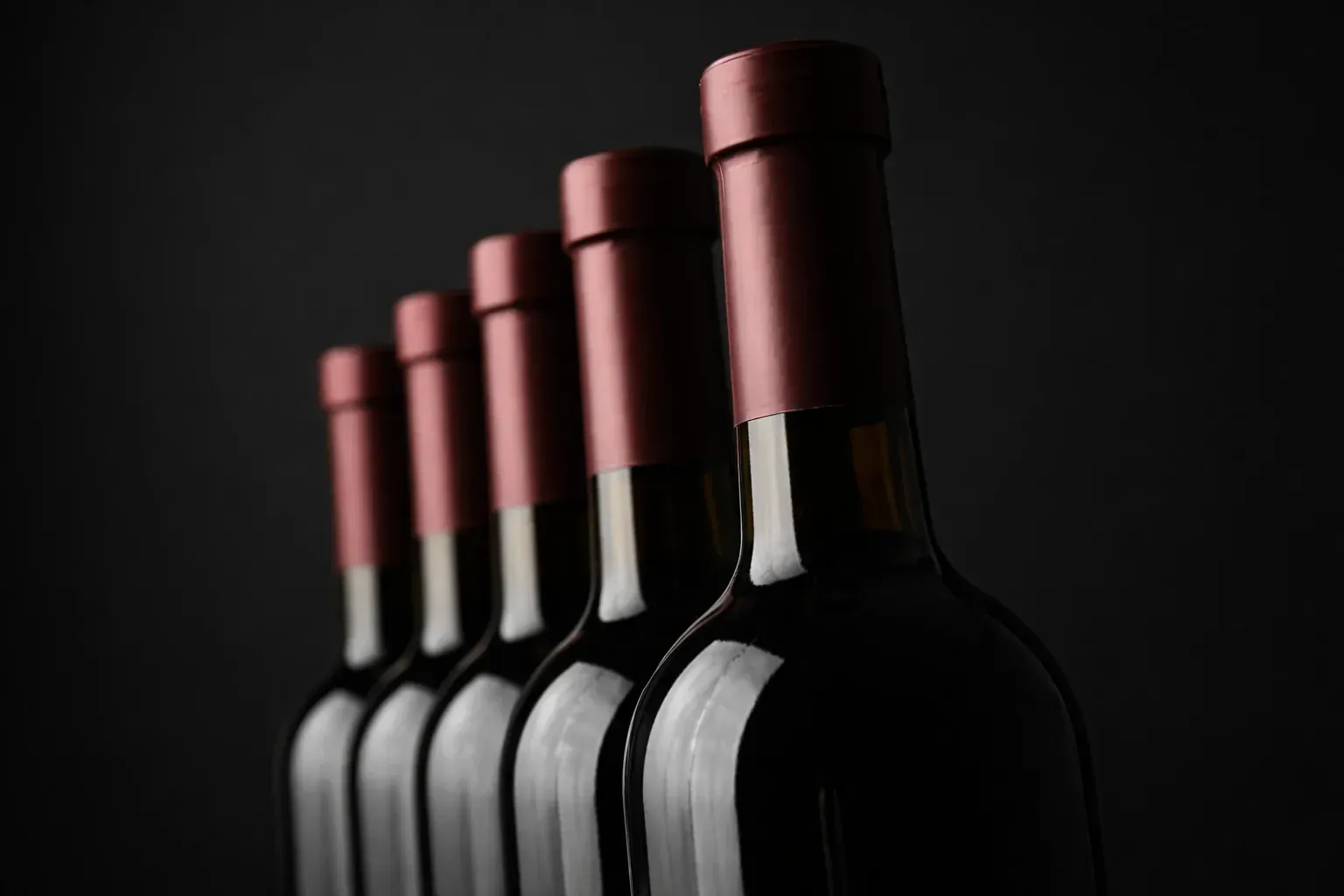 Cinco botellas de vino oscuro con tapones de papel de aluminio color granate se alinean en diagonal sobre un fondo negro.