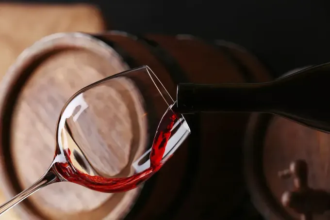El vino tinto se vierte de una botella a una copa frente a barricas de madera.