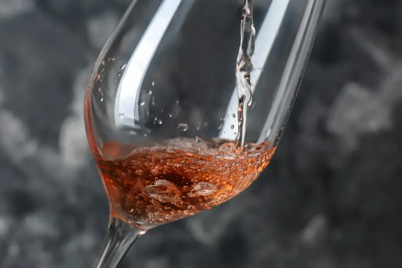 Vino rosado vertiéndose en una copa con tallo sobre un fondo oscuro y texturizado.