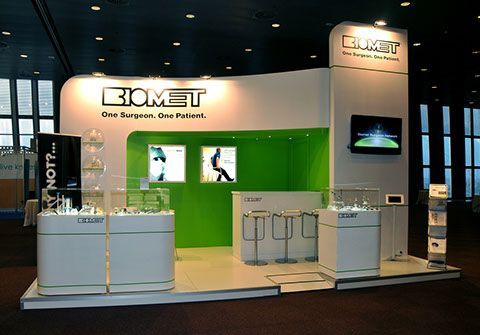 Stand de Biomet en la feria con detalles en blanco y verde. Presenta productos, señalización y una pantalla de televisión.