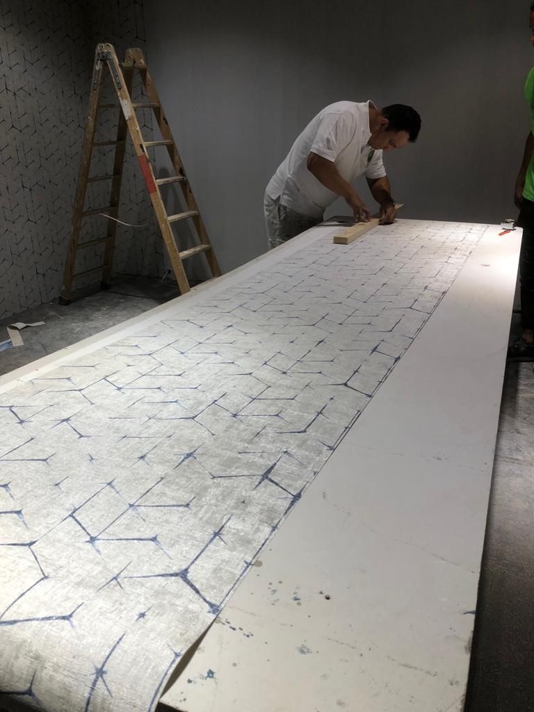 Un hombre corta papel tapiz sobre una mesa; escalera y pared gris en el fondo.