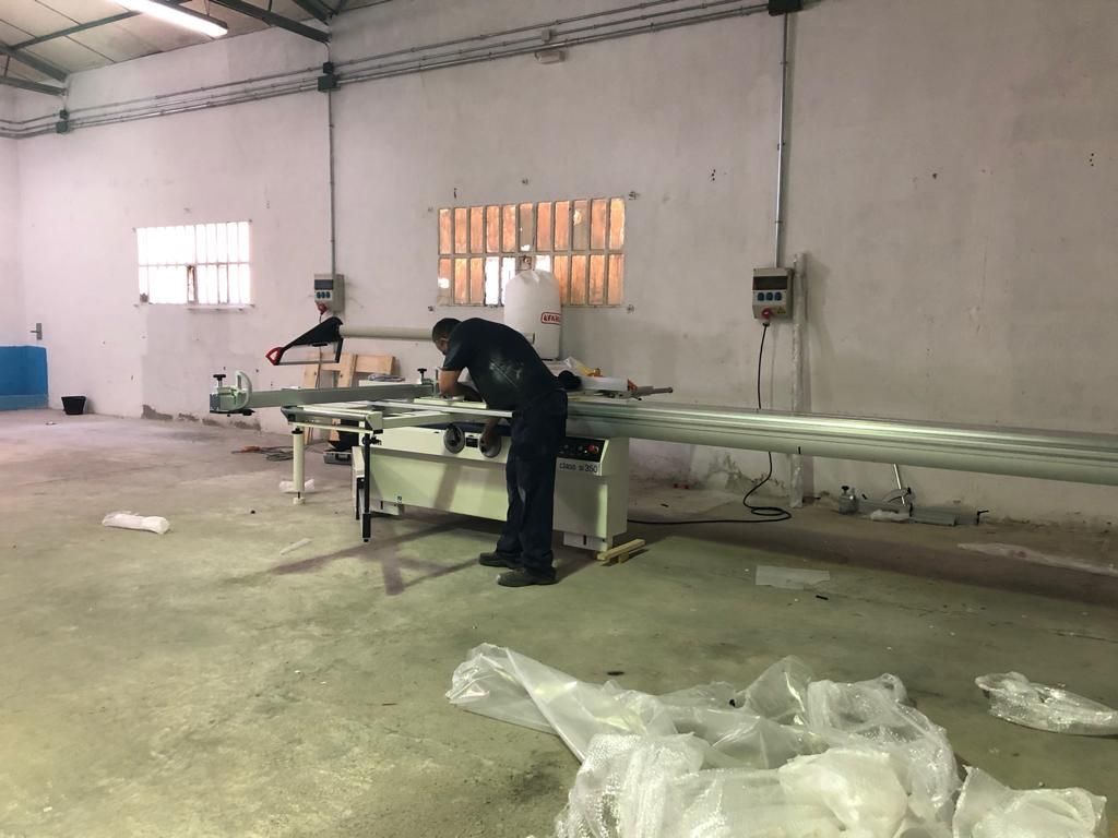 Hombre utilizando una sierra en un taller con un largo trozo de metal, un interior luminoso con suelo de hormigón.