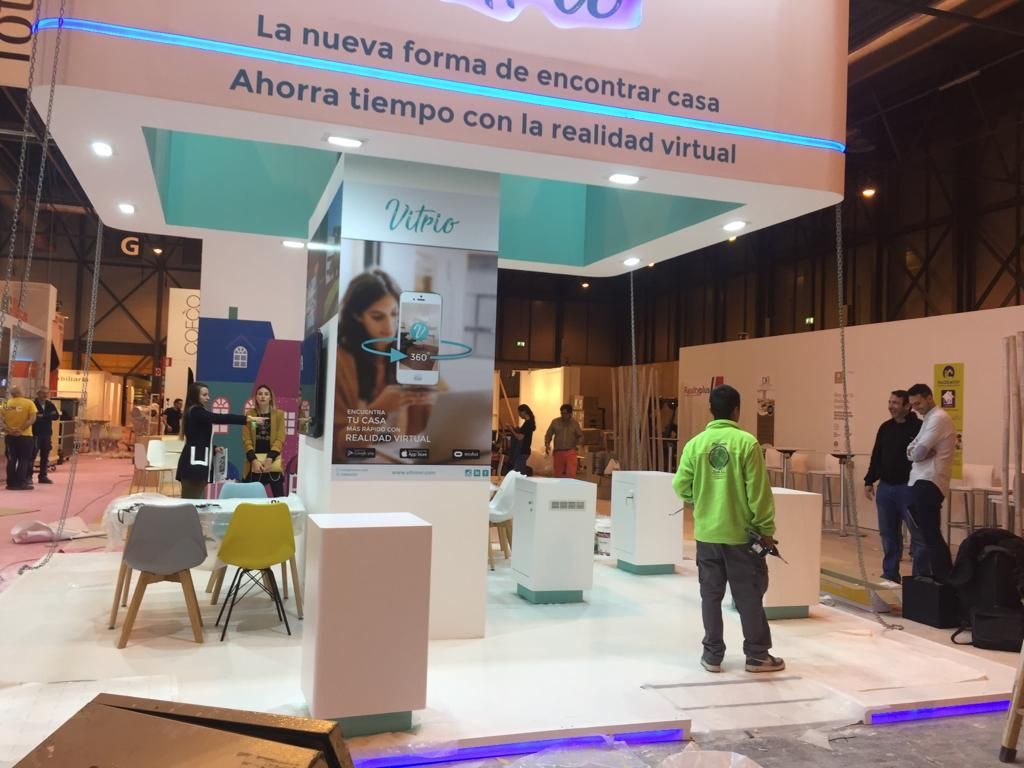 Stand de feria con estructuras blancas y gente instalándose. Un cartel turquesa y blanco dice 