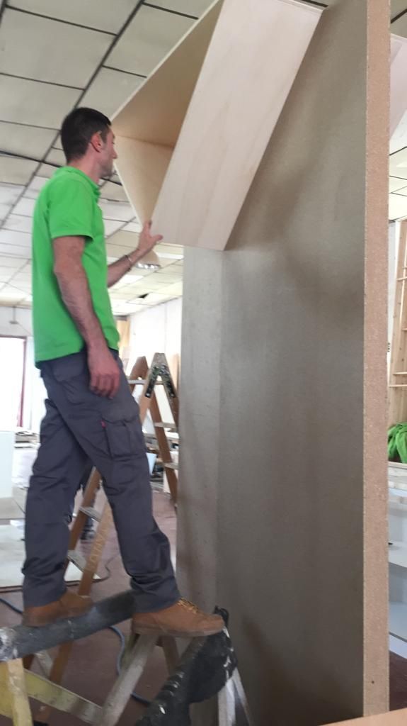 Hombre con camisa verde en una escalera de mano instalando un estante de madera en una gran pared divisoria.