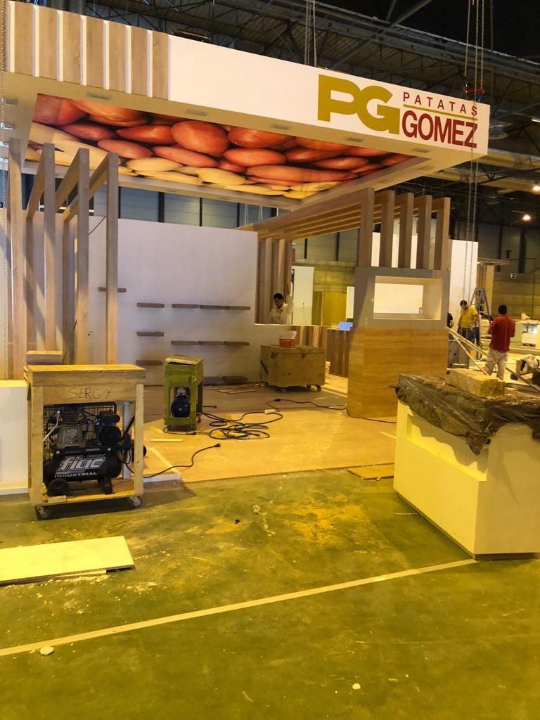 Stand de feria en construcción con letreros de PG Palate Gomez. Trabajadores, madera y equipo visibles.