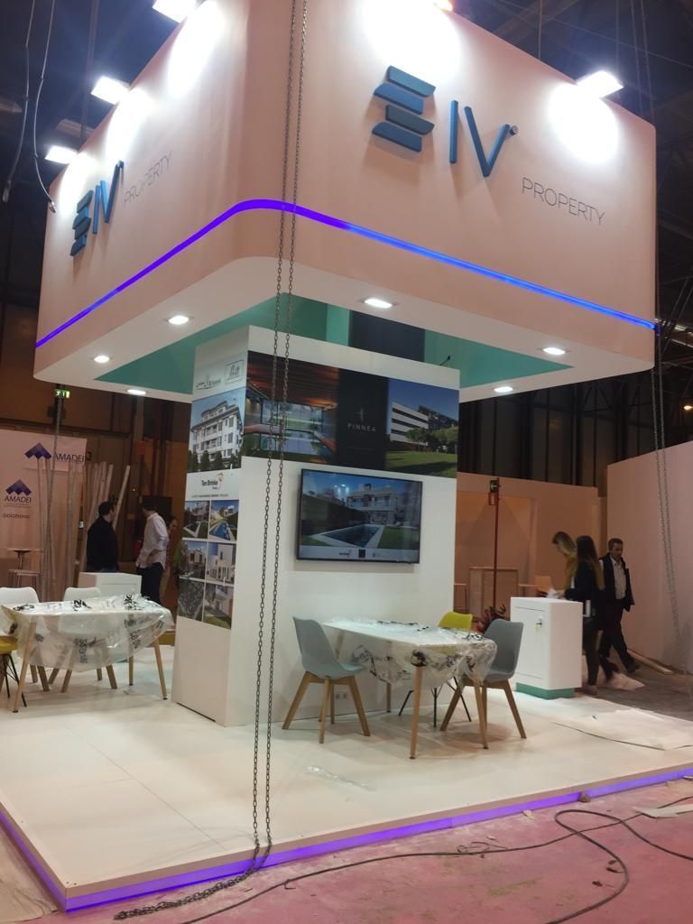 Stand de feria: logo “IV”, diseño en blanco y turquesa, pantallas, mesas y sillas, gente al fondo.