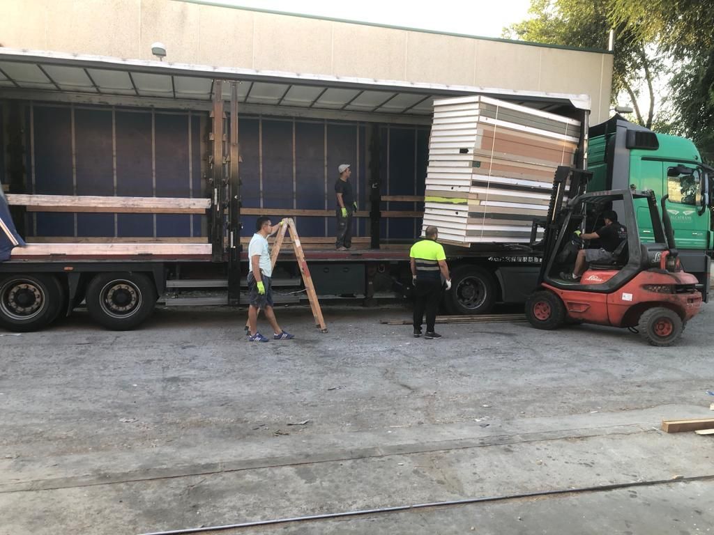 Trabajadores descargando paneles de un camión con una carretilla elevadora. Exterior, suelo de grava gris.