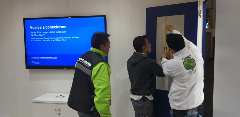 Tres personas instalando algo en una puerta azul. Una mide, otra observa. La pantalla del televisor es azul.