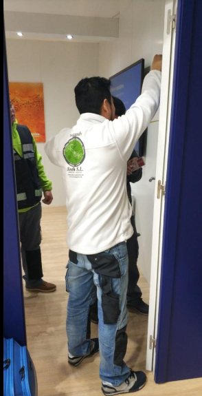 Una persona con camisa blanca y jeans instala algo en una pared mientras otra persona observa.