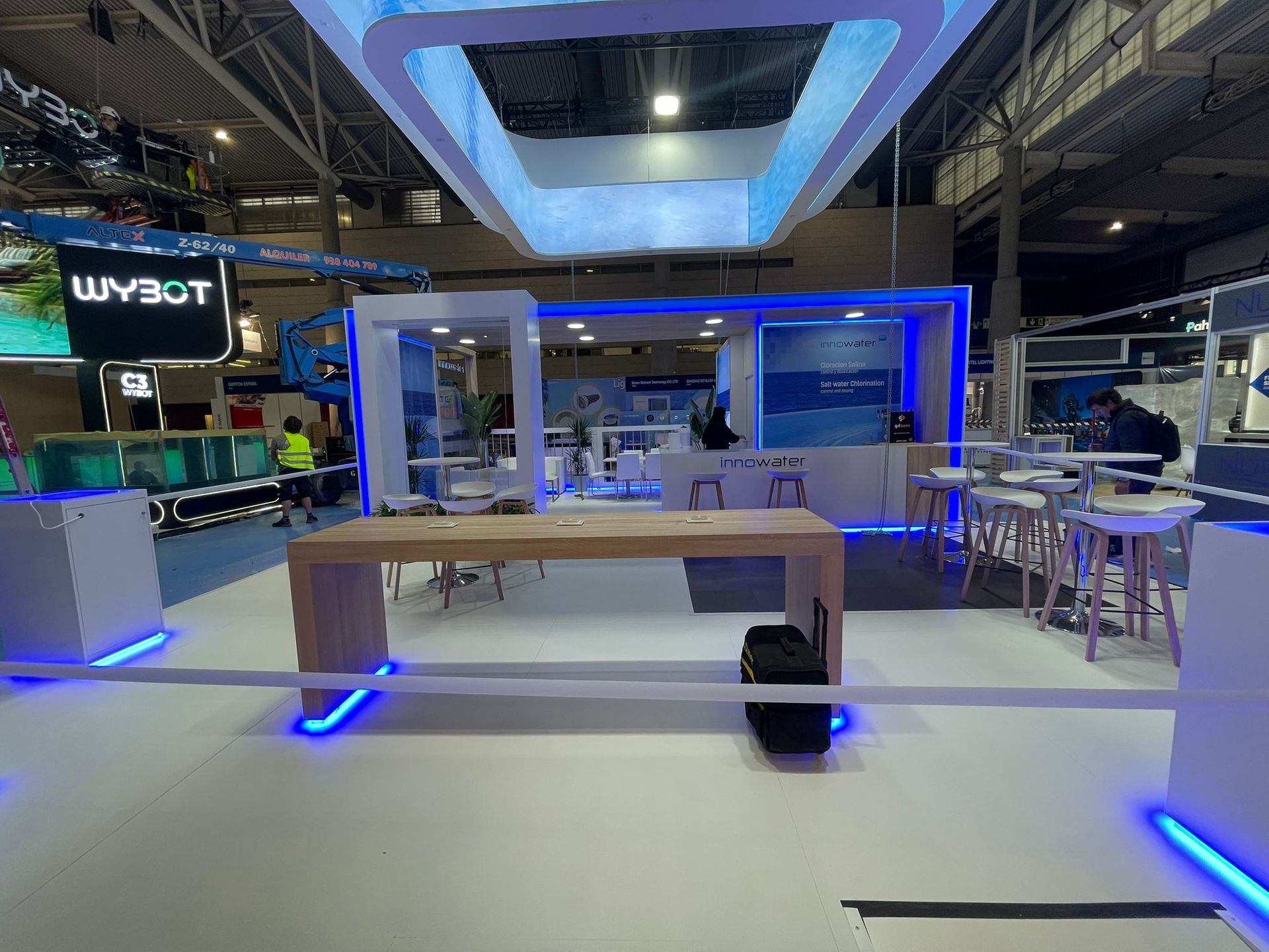 Stand de exposición con iluminación azul, mesa de madera clara y taburetes. Se ven personas al fondo.