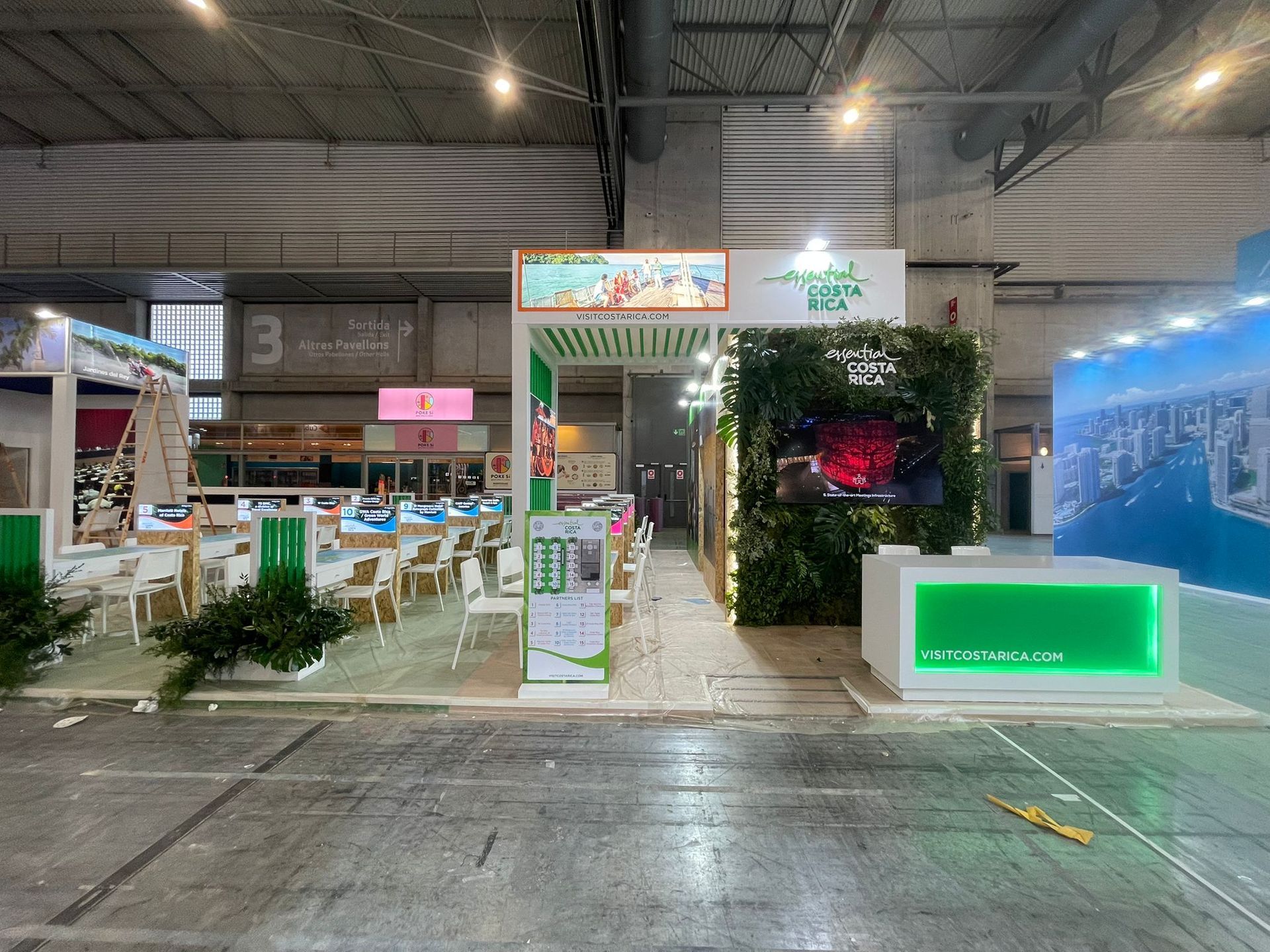 Un stand de feria comercial con detalles en verde y blanco, que cuenta con una pantalla y una zona de asientos.