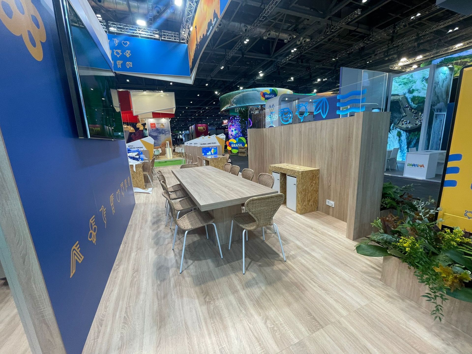 Interior de un stand de feria comercial con una mesa larga, sillas y paneles de madera.